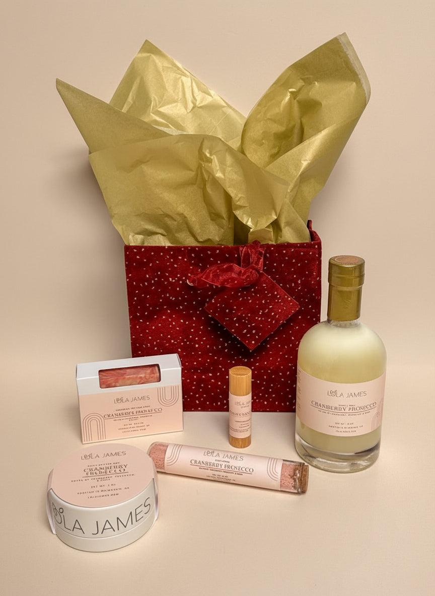 Skincare products and a bottle of bubble bath in a decorative burgundy bag with gold tissue paper on a neutral background.
