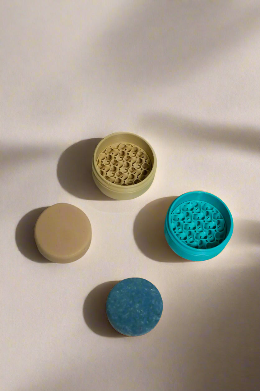 Blue shampoo bar with blue travel case, beige conditioner bar with beige travel case on a neutral background.