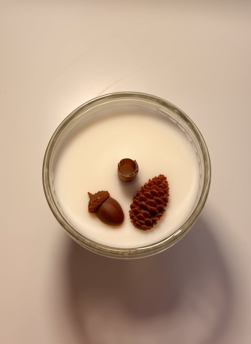 Candle in a glass jar with decorative acorn and pine cone designs on a neutral background