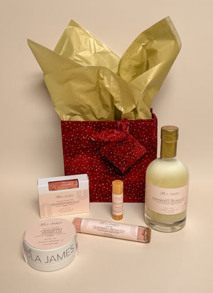 Skincare products and a bottle of bubble bath in a decorative burgundy bag with gold tissue paper on a neutral background.