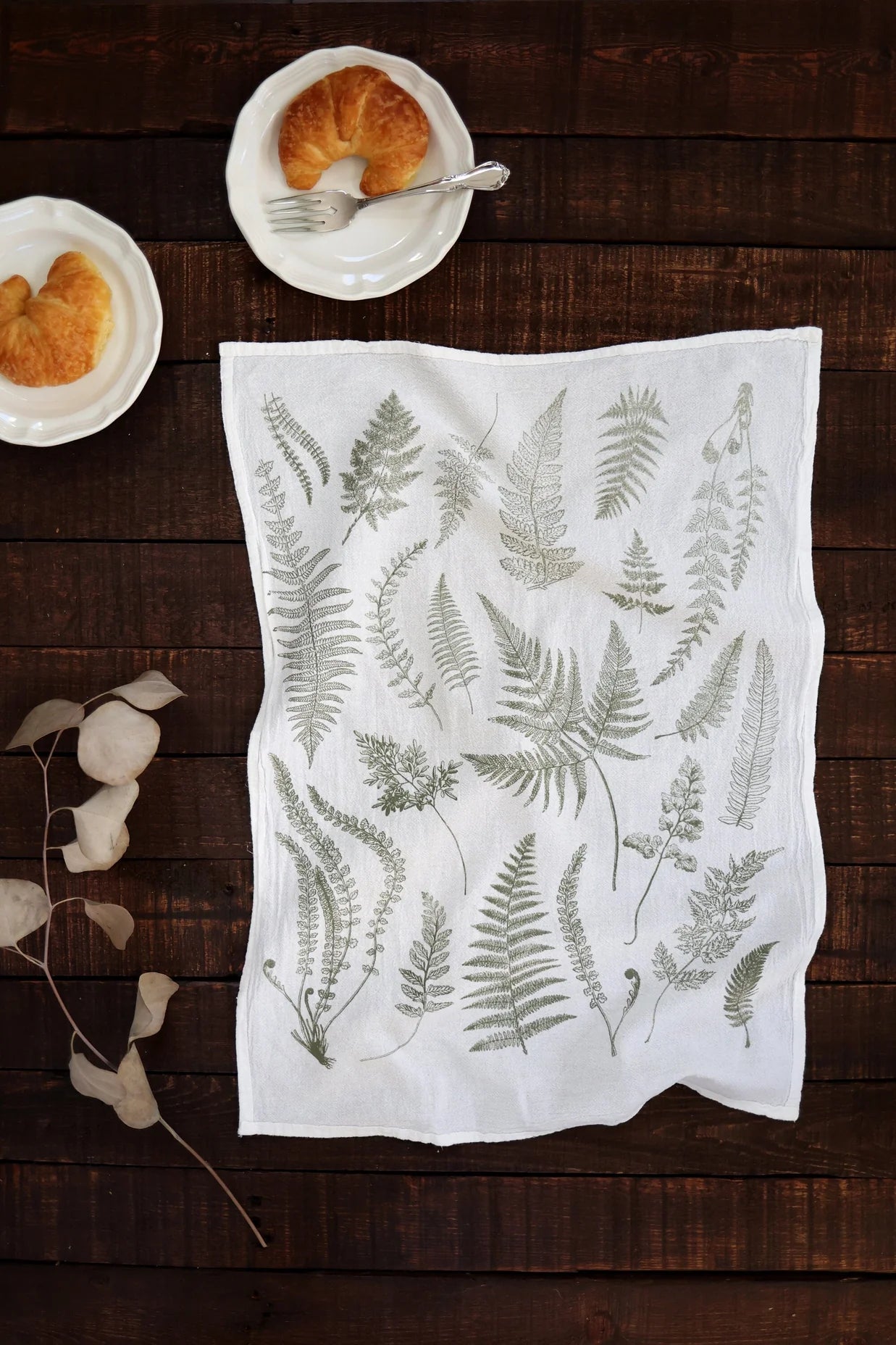Tea towel with fern pattern on a wooden table with pastries and plates.